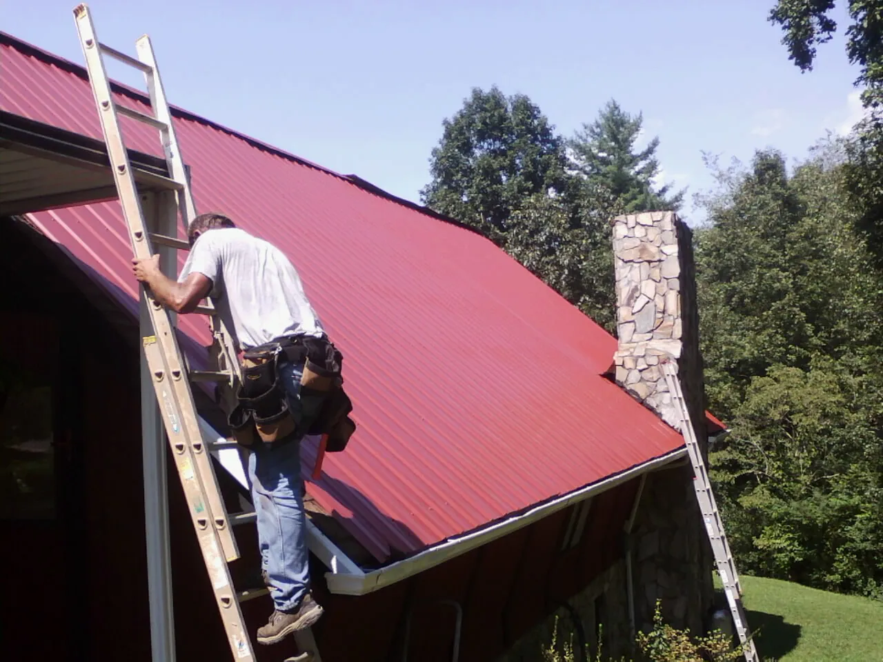Roofer on a ladder beside a red metal roof and stone chimney — Soffit & Fascia Repair by Roofing Brothers in Saint Thomas